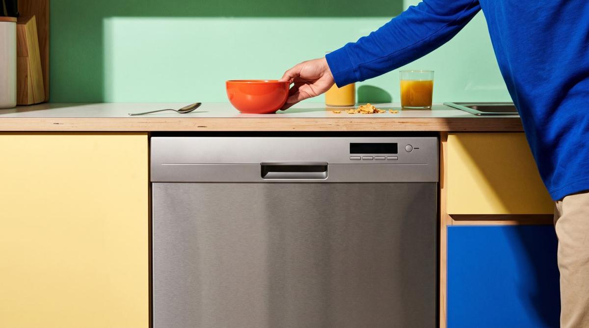 A stainless steel dishwasher in a sunny kitchen with mint walls, yellow cabinets, and blue accents