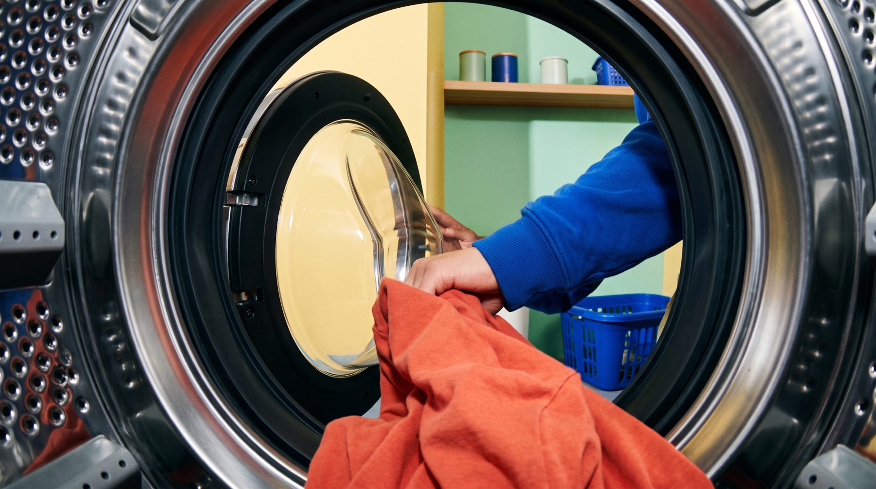 Hands loading orange laundry, viewed from inside the washing machine drum